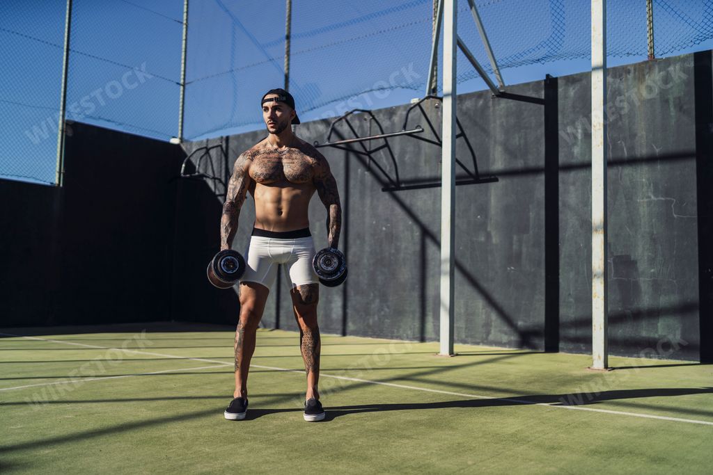 An attractive tattooed man working out with dumbbells in an outdoor court on a sunny day