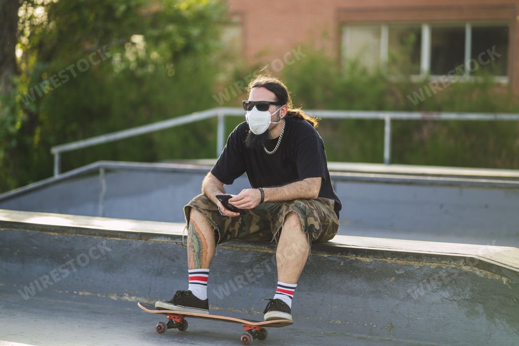 A young male sitting with a skateboard in the park wearing a medical face mask - Covid-19 concept