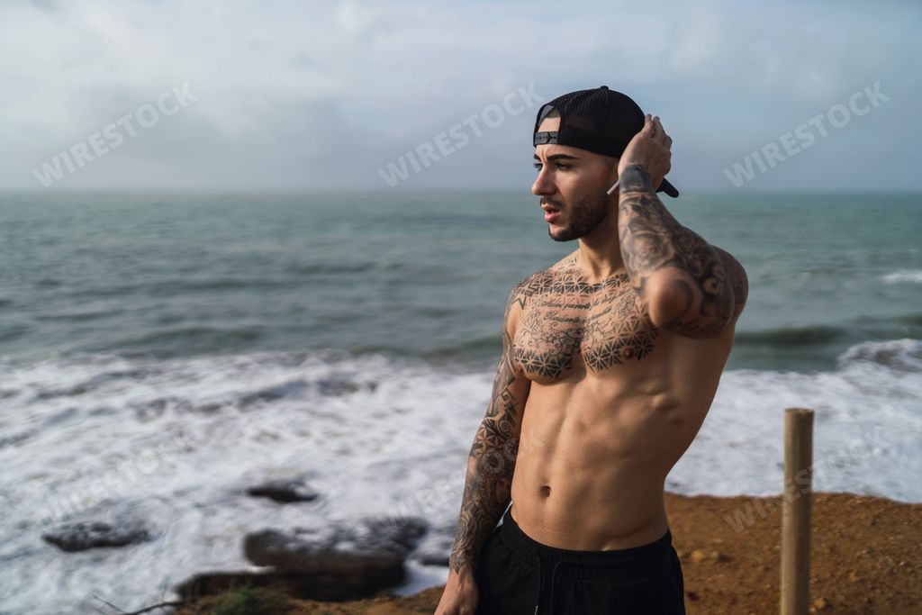 A topless Caucasian man from Spain with tattoos posing in a beach