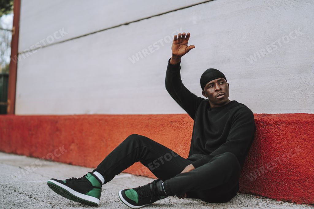 A sporty Black man in black sweaters and jogging pants sitting on concrete pavement outside a building