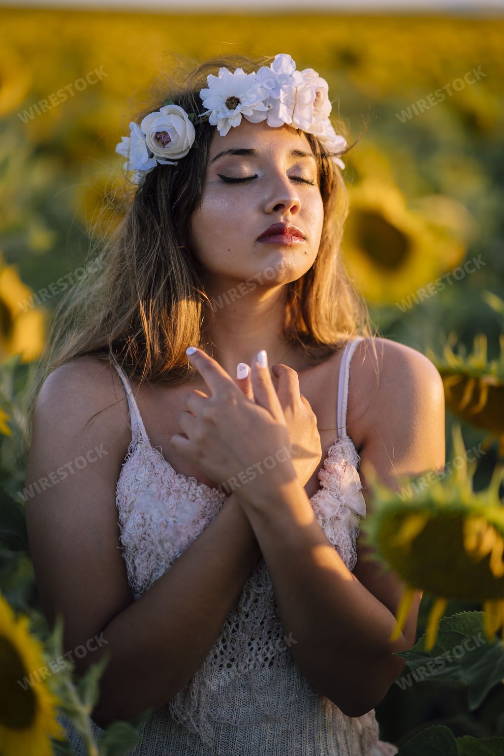 A vertical portrait of a young brunette female  in a flower crown with closed eyes holding clasped hands up to her chest in a sunflower field
