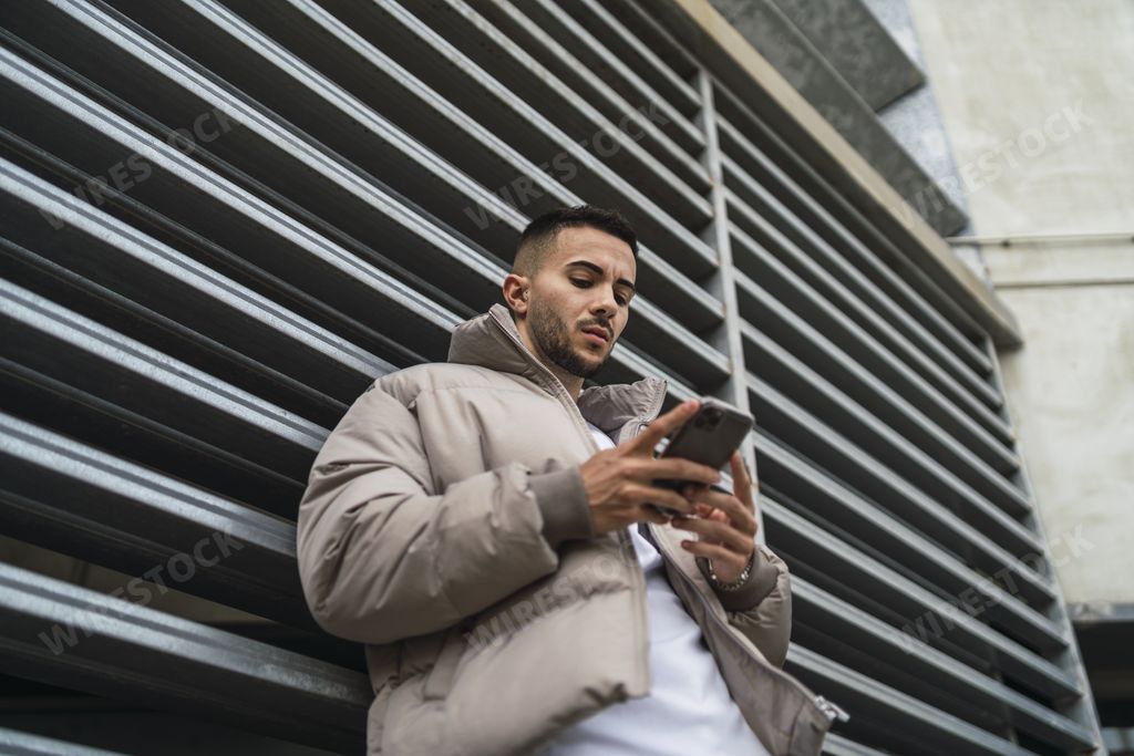 A low angle shot of a sexy male in a street style outfit using his phone outdoors