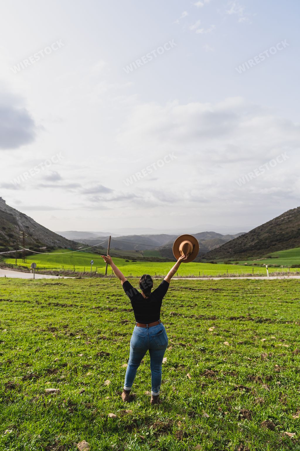 A back shot of an excited adult woman standing in the field looking at the mountains and cloudy sky on a sunny day