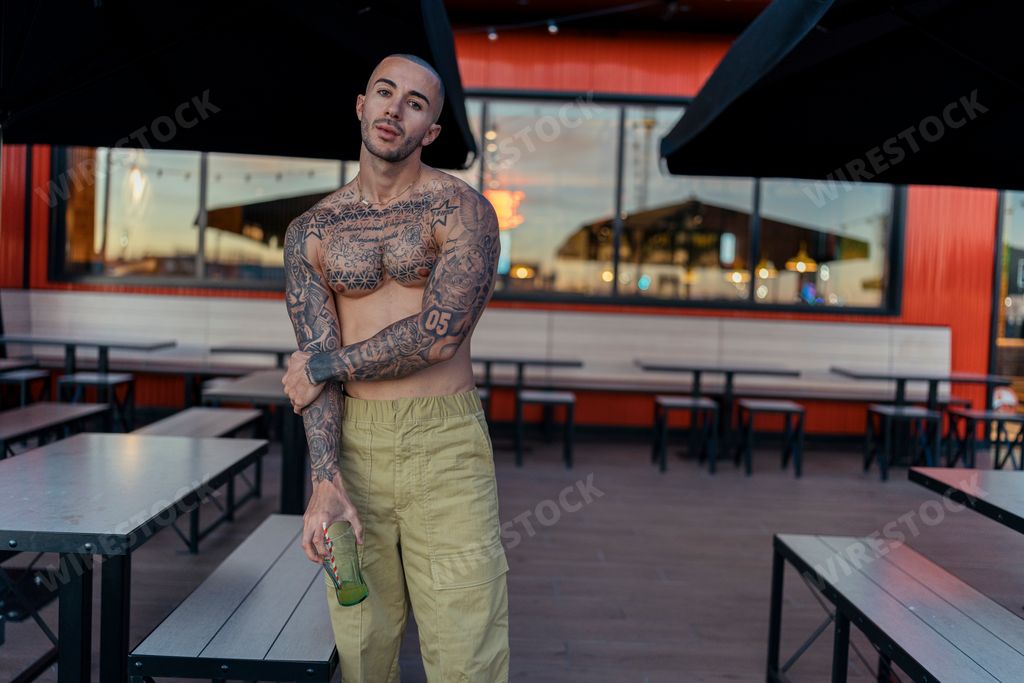 A handsome shirtless man with tattoos, posing against an outdoor restaurant