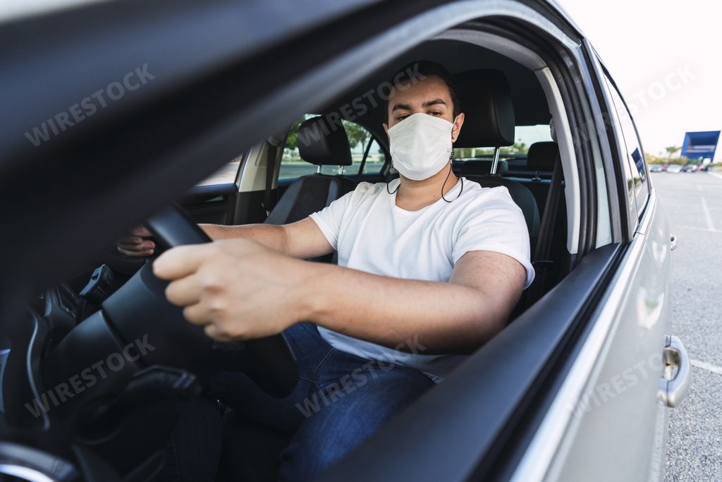 A young Caucasian male driving a car with a medical face mask - the new normal after Covid-19