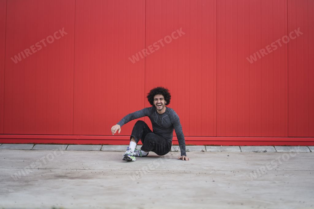 A laughing stylish Spanish guy with curly hair sitting on the ground in the red wall background