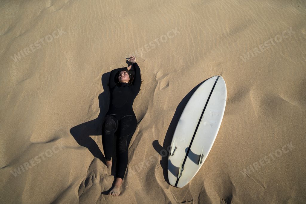 An aerial shot of an attractive female lying down on a sandy ground with a surfboard on the side