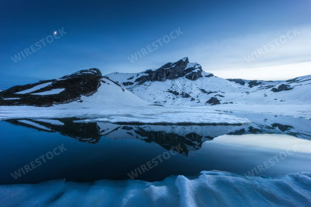 A scenic view of a snow-capped mountain reflecting in a partially frozen lake at Etang de Trente Pas, Wallis, Switzerland