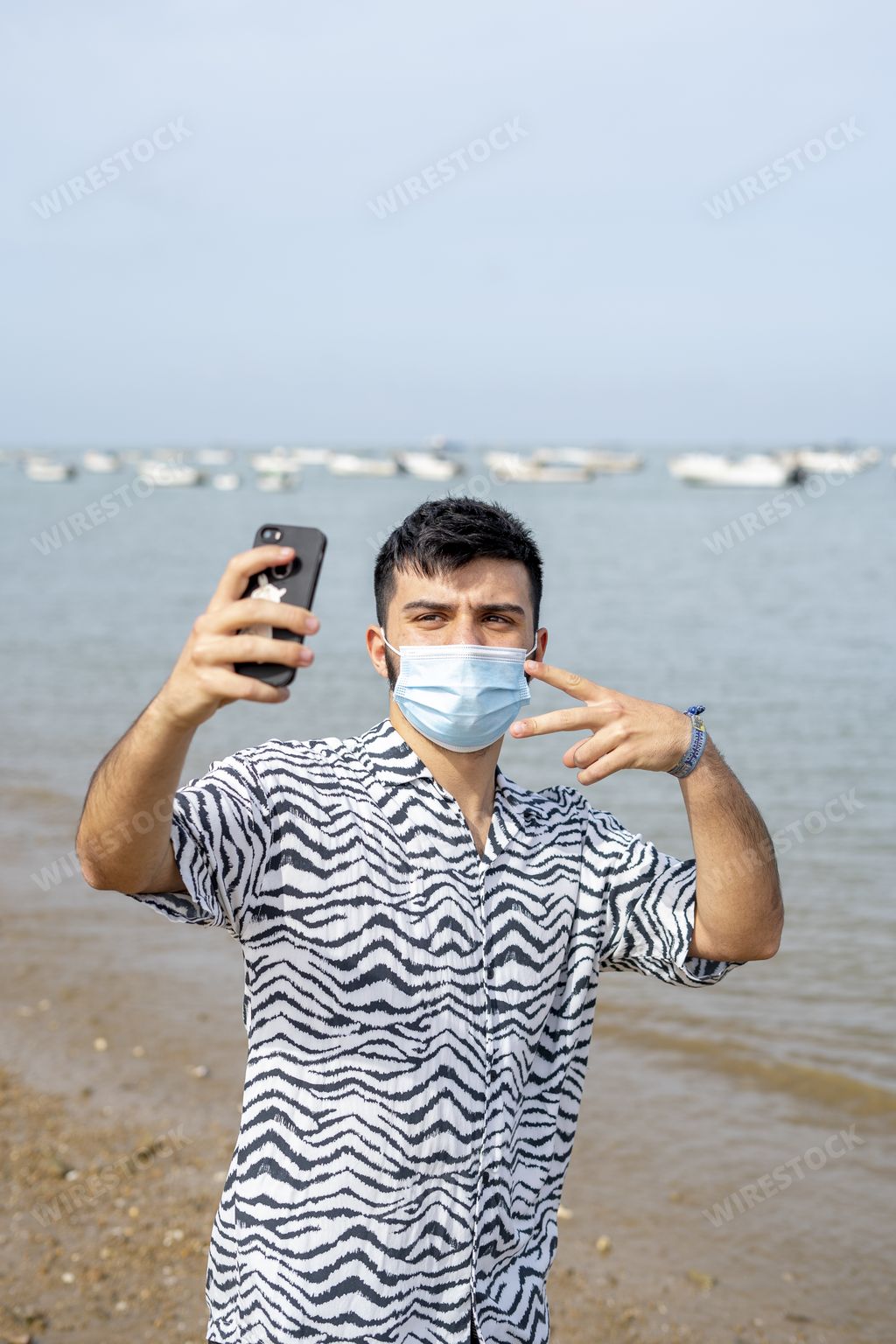 A Hispanic man wearing protective mask while taking selfies with peace hand sign at a beach