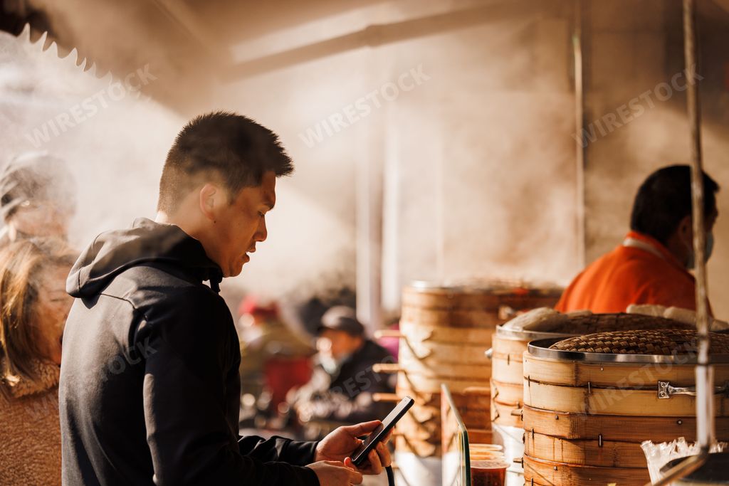 A closeup of a handsome Asian man looking at his phone at a steamed dumpling shop at sunset