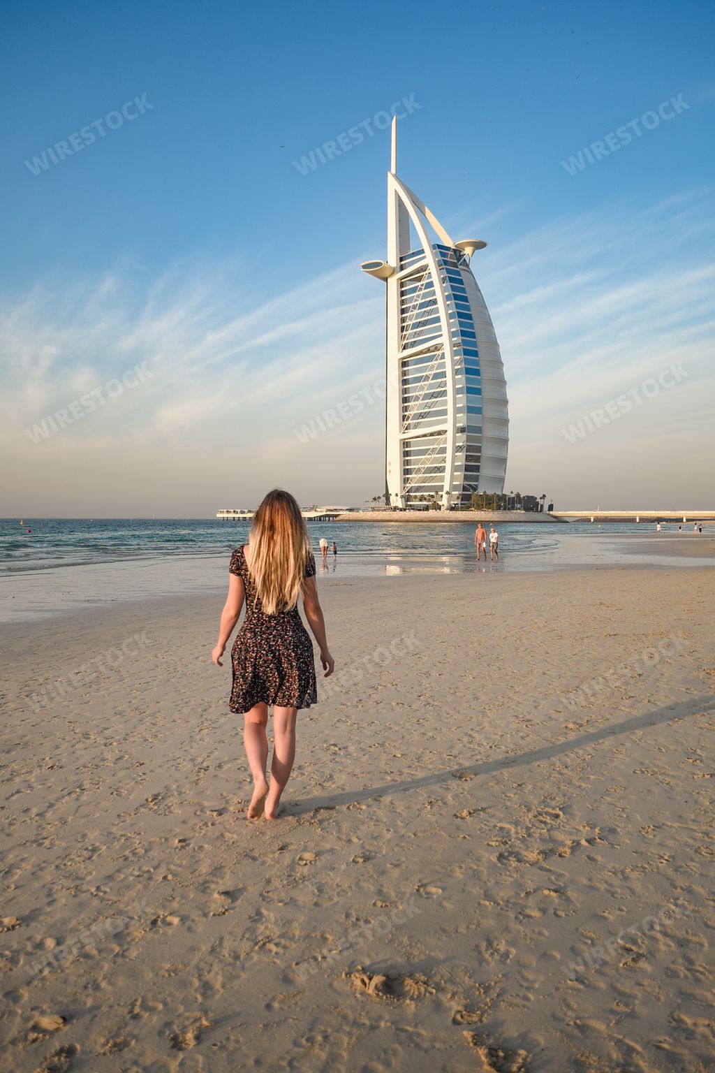 A back view of a young blonde female on Jumeirah Beach and luxury Burj al Arab hotel in background, Dubai, United Arab Emirates