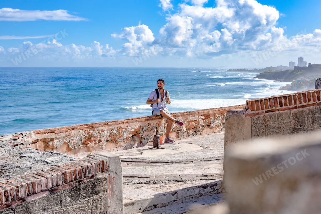 An attractive sexy Hispanic male sitting on a parapet wall having a beautiful wavy seascape behind