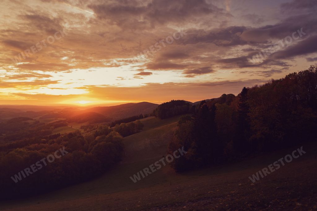 A breathtaking view of a sunset from the top of a hill, surrounded by lush hillsides dotted with trees
